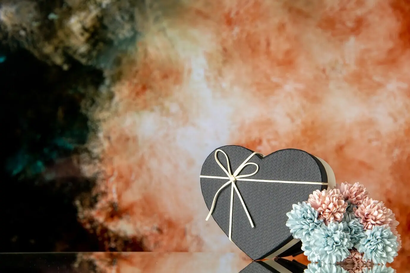 Vorderansicht einer herzförmigen Geschenkbox mit schwarzem Deckel und farbigen Blumen vor abstraktem schwarz-beigem Hintergrund
